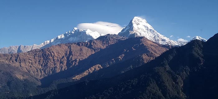 "Image of trekkers standing on Poon Hill at sunrise, surrounded by majestic Himalayan peaks, including Annapurna and Dhaulagiri. The sky is painted in hues of orange and pink, and the trekkers are taking in the breathtaking views of the Himalayan range. The surrounding landscape is covered in lush forests, and the village of Ghorepani can be seen in the distance."