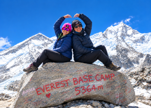 Couple sitting on Everest Base Camp rock with snowy mountain views.