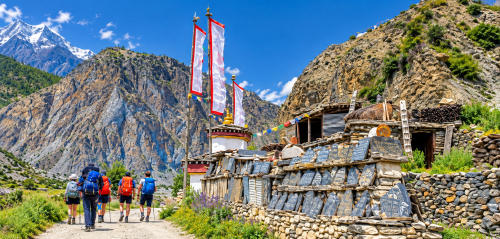 Trekkers walking past a traditional stone Mani wall, colourful Buddhist prayer flags and a mountain village along the Annapurna Circuit Trek in Nepal.