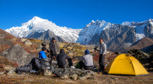 Langtang Ganjala Pass Trek campsite with trekkers relaxing beside a yellow tent under clear blue skies, surrounded by snow-covered Himalayan peaks in Langtang National Park, Nepal.