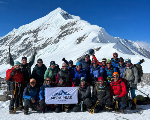 Group of international climbers and Sherpa guides celebrating a successful summit on Mera Peak, standing on snowy high-altitude terrain with full mountaineering gear, smiling together in front of a dramatic Himalayan peak under a clear blue sky- Mera Peak Climbing
