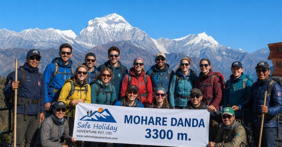 Mohare Danda Trek- Group of trekkers and Nepali guides posing at Mohare Danda viewpoint with Himalayan scenery, including Mount Dhaulagiri in the background, green hills, and a banner reading “Safe Holiday Adventure,” under a clear blue sky.