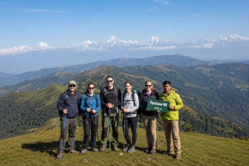 Group of hikers with a local Nepali guide standing on the summit of Panchase Hill (2,500 m) near Pokhara, Nepal, surrounded by rolling green hills and panoramic Himalayan mountain views under a clear blue sky.