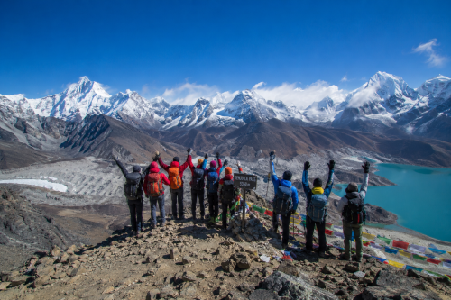Group of trekkers standing at the summit of Renjo La Pass with panoramic Himalayan mountain views and Gokyo lakes below.
