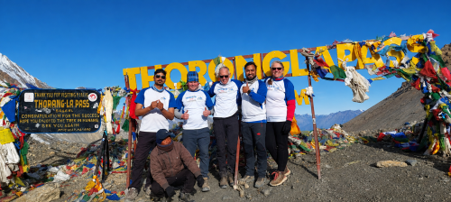 A group of six people posing at Thorong La Pass (5,416 m) on the Annapurna Circuit Trek under a bright blue sky. Colorful Buddhist prayer flags surround the iconic yellow Thorong La Pass sign, with rugged Himalayan mountains and rocky alpine terrain creating a spectacular high-altitude backdrop.