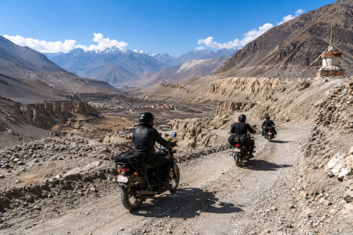 A group of adventure riders on Royal Enfield motorcycles journey through the dramatic landscapes of Upper Mustang, Nepal. The riders travel along a dusty dirt road near Kagbeni Valley under a clear blue sky, surrounded by colorful cliffs, barren hills, and ancient Tibetan-style villages. Snow-capped Himalayan peaks rise in the distance, while prayer flags and rugged canyon scenery showcase the remote beauty of the Forbidden Kingdom. The image captures the spirit of an Upper Mustang motorbike tour, blending culture, adventure, and spectacular mountain landscapes.