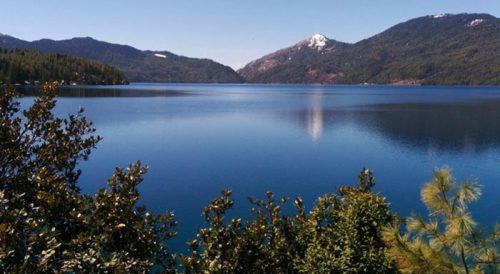 A serene view of Rara Lake surrounded by lush green hills and mountains, reflecting clear blue water under a calm sky.