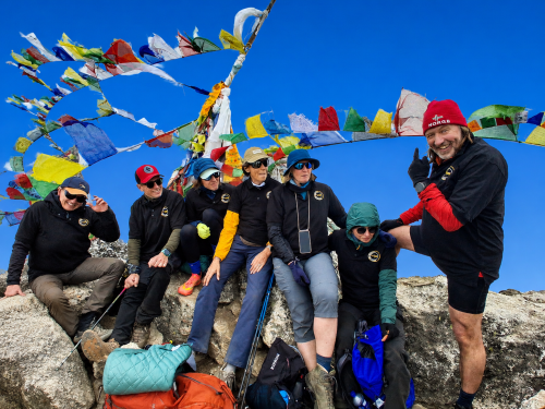 Trekkers posing with colorful prayer flags during the Everest High Pass Trek in the Himalayas beneath a clear blue sky.